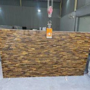 May include: Large rectangular slab of brown and gold stone with a textured, brick-like pattern. The stone is suspended by a crane hook in a warehouse setting. The stone appears to be a countertop or decorative panel.