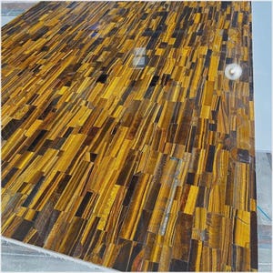 May include: A large, rectangular surface composed of tiger's eye gemstone pieces. The surface has a mosaic-like appearance with varying shades of gold, brown, and black. The pieces are arranged in a linear pattern, creating a textured and visually striking design.