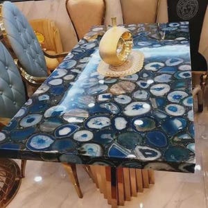 May include: A rectangular dining table featuring a polished surface composed of numerous blue and white agate slices embedded in resin. The table is supported by a gold-toned metal base and holds a gold-colored decorative bowl on a lace doily.