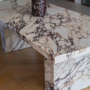 Puede incluir: Una mesa de centro rectangular de mármol con un patrón veteado blanco y marrón. La mesa tiene una base sólida y una tapa a juego. Un jarrón marrón con flores está sobre la mesa. La mesa es de mármol.