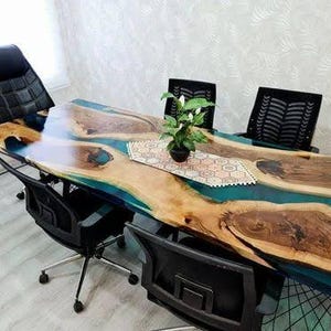 May include: A large, custom-made wooden table with a blue epoxy river design. The table is surrounded by black office chairs. A small potted plant sits on a decorative runner in the center of the table.