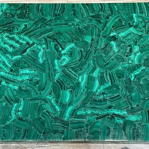 Puede incluir: Losa rectangular de piedra de malaquita con superficie pulida. La piedra presenta un patrón arremolinado de tonos verdes oscuros y claros, creando un diseño natural y decorativo. La losa es adecuada para diversas aplicaciones decorativas.