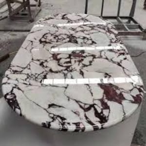 May include: Oval marble table with a white base and a polished top. The tabletop features a striking pattern of white, grey, and burgundy veining. The table is approximately 152 cm long and 91 cm wide.