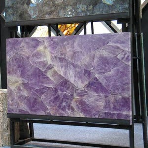 May include: A large rectangular amethyst slab with a mosaic pattern of purple and white crystalline formations. The slab is mounted on a black metal frame, showcasing the natural beauty of the gemstone. The amethyst is a deep purple color.