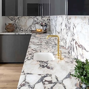 Puede incluir: Una cocina moderna con encimera de mármol blanco con vetas grises oscuras. Se instala un grifo dorado y un fregadero blanco. También se ven armarios grises oscuros y un suelo de madera. Un jarrón, pan y botellas de vino están en la encimera.
