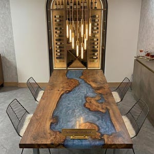 May include: A rectangular dining table featuring a natural wood slab with a central blue resin river inlay. The table has metal legs and is surrounded by four black wire-frame chairs with white cushions. A gold plaque reads TheHandiCraftsHubCo.