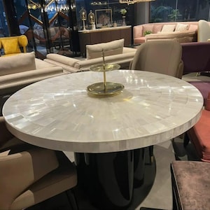 May include: A circular dining table with a white, mosaic-style top and a black pedestal base. A two-tiered gold serving tray is on the table. The table is surrounded by chairs and sofas in a showroom.