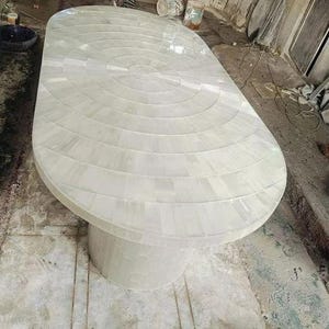 Könnte beinhalten: Ein ovaler Tisch mit einer weißen, mosaikartigen Oberfläche. Die Tischplatte weist ein konzentrisches kreisförmiges Muster auf. Der Tisch steht auf einem soliden, zylindrischen Sockel. Das Design ist modern und elegant.