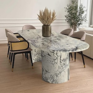 Pode incluir: Mesa de jantar oval de mármore com base cilíndrica e seis cadeiras estofadas. As cadeiras têm pernas pretas com pontas douradas e encostos de palhinha tecida. Um vaso com plantas secas está sobre a mesa.