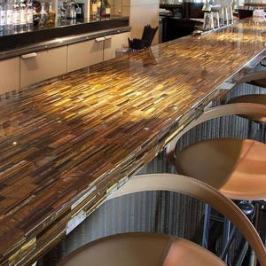 May include: A long, rectangular bar with a polished, mosaic-like surface in shades of brown and gold. The bar is lined with several tan leather and chrome bar stools. The bar is in a restaurant or lounge setting.