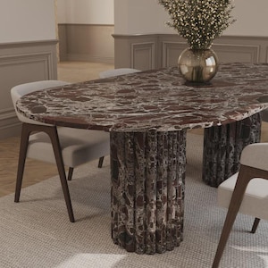May include: Oval-shaped dining table with a red and brown marble top and two cylindrical, textured bases. The table is surrounded by light gray chairs with dark wooden legs. A vase of flowers sits on the table.