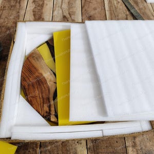 May include: A circular wooden board with a natural grain pattern and a yellow resin inlay, partially covered by white protective foam sheets inside a cardboard shipping box. The item features a smooth finish and a distinct contrast between the wood.