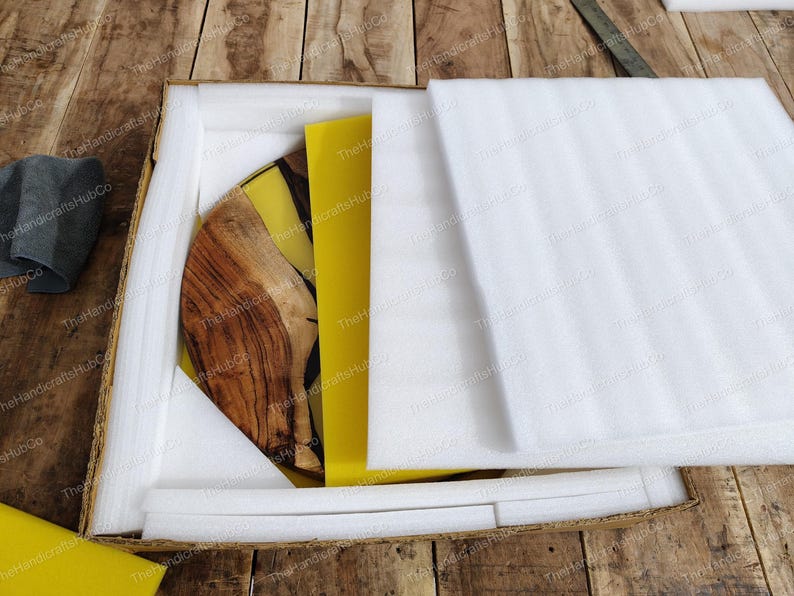 May include: A circular wooden board with a natural grain pattern and a yellow resin inlay, partially covered by white protective foam sheets inside a cardboard shipping box. The item features a smooth finish and a distinct contrast between the wood.