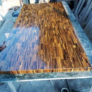 May include: A large, rectangular wooden tabletop with a mosaic-like pattern. The surface is composed of numerous small, rectangular pieces of wood in shades of brown, gold, and black. The wood has a glossy finish, reflecting light and creating a visually striking effect.