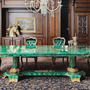Puede incluir: Un comedor lujoso con una gran mesa rectangular de malaquita con detalles dorados. La mesa está rodeada de sillas ornamentadas con respaldos de terciopelo verde. La habitación tiene paneles de madera oscura y una lámpara de araña.