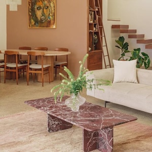 May include: A rectangular red marble coffee table with white veining. The table is in a living room with a beige sofa, a rug, and a vase of greenery. A dining table and chairs are visible in the background.