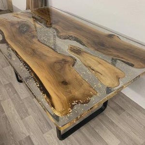Op de afbeelding: Een rechthoekige houten tafel met een helder epoxyharsblad. De tafel heeft natuurlijke houtnerfpatronen en een zwart metalen onderstel. De tafel is ongeveer 152 cm lang en 76 cm breed.