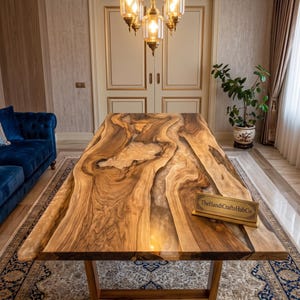 Op de afbeelding: Een rechthoekige eettafel met een natuurlijk houten blad met boomstamrand en een centrale inleg van doorschijnende hars. De tafel heeft een gladde, gepolijste afwerking en een houten onderstel. Een klein goudkleurig bordje toont TheHandiCraftsHubCo.