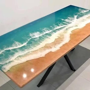May include: A rectangular table with a resin top depicting a beach scene. The top features a gradient of colors, transitioning from turquoise to tan, with white wave details. The table has black metal legs.