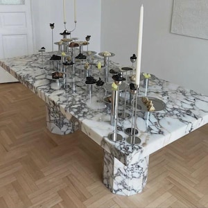 Puede incluir: Una gran mesa de comedor rectangular de mármol con un patrón veteado blanco y gris. La mesa está sostenida por dos bases de pedestal geométricas. En la mesa se disponen fuentes de plata y candelabros con velas encendidas, mostrando pequeños alimentos.