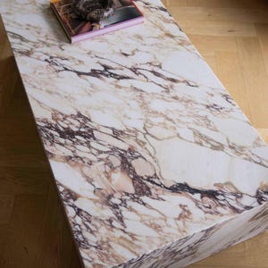 Op de afbeelding: Een rechthoekige marmeren salontafel met een wit en bruin geaderd patroon. Een boek en een donkergekleurd decoratief object liggen op het oppervlak van de tafel. De tafel staat op een houten vloer.