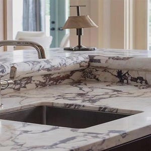 May include: A kitchen countertop featuring a stainless steel sink and tap. The countertop is white with grey and brown veining. A small lamp with a beige shade sits on the countertop.