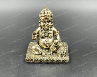 Può includere: Una statua in ottone dettagliata di una divinità, seduta su una base rettangolare decorativa. La figura indossa una corona, una collana e altri ornamenti. Un piccolo vaso è posto a destra. La statua ha un'espressione sorridente.
