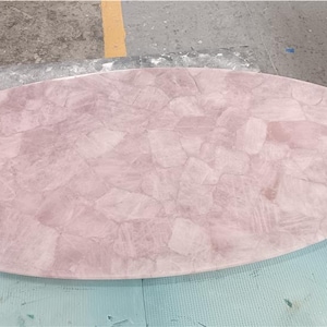 May include: Oval-shaped rose quartz table top. The surface is a mosaic of pink and white stone fragments, creating a unique, textured appearance. The table top is approximately 60 inches long.