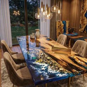 Pode incluir: Uma mesa de jantar retangular com um tampo de madeira de borda natural e uma incrustação de resina azul. A seção de resina retrata uma cena de praia com seixos, ondas de espuma branca e estrelas do mar. Uma placa dourada diz TheHandiCraftsHubCo.