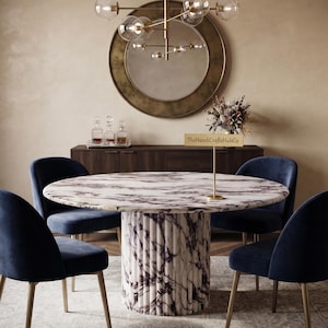 May include: A round dining table with a white and purple marble top and a fluted marble base, surrounded by four navy blue velvet chairs with gold-toned legs. A gold sign on the table displays the text TheHandiCraftsHubCo in black lettering.