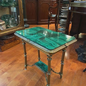 Puede incluir: Una mesa auxiliar antigua con tapa rectangular de malaquita y marco de latón. La mesa tiene detalles ornamentados en las patas y un estante inferior. La tapa de malaquita tiene un patrón verde arremolinado. La mesa mide aproximadamente 61 cm de largo, 41 cm de ancho y 71 cm de alto.