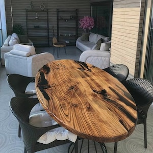 Op de afbeelding: Een ovale houten tafel met een natuurlijke houtnerf afwerking en donkere accenten. De tafel is omringd door zwarte rieten stoelen, waardoor een eethoek of zithoek buiten ontstaat. De kamer heeft meubels en decoraties in neutrale tinten.