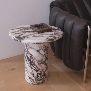 Puede incluir: Una mesa auxiliar redonda de mármol con base cilíndrica. La mesa tiene una parte superior y una base con un patrón blanco y marrón. Un cenicero de mármol marrón está sobre la mesa. La mesa está junto a una silla de cuero negro.