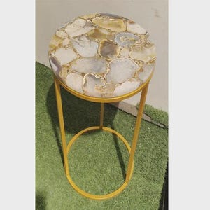May include: A small, round side table with a natural stone top. The tabletop features a mosaic of white, grey, and gold-toned stones. The table has a slender, gold-coloured metal frame, with a circular base for stability.