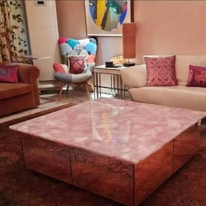 May include: A large, square coffee table with a pink, patterned top and mirrored sides. The table is the focal point of a living room with a brown sofa, patterned chair, and a white sofa with decorative pillows.
