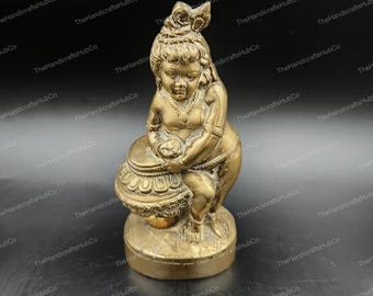 Può includere: Una statua color oro di una figura seduta che tiene in mano un oggetto rotondo. La figura ha lineamenti dettagliati ed è adornata da un copricapo e gioielli. La statua è su una base circolare.
