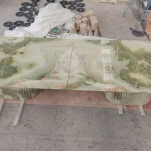 Op de afbeelding: Een grote, rechthoekige tafel met een gepolijst, lichtgroen en wit marmeren blad. Het marmer heeft donkergroene adering. De tafel wordt ondersteund door twee cilindrische bases en een houten frame. De tafel is ongeveer 183 cm lang.