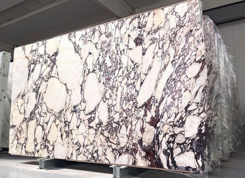 May include: Large slab of marble with a striking pattern of white, purple, and black veining. The stone is displayed on a metal support, showcasing its size and natural beauty. The marble has a polished surface.