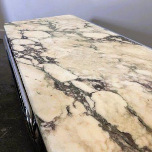 May include: Close-up of a rectangular table top with a polished marble surface. The marble features a cream-coloured base with dark grey and purple veining. The table has a shiny, chrome-coloured metal trim.