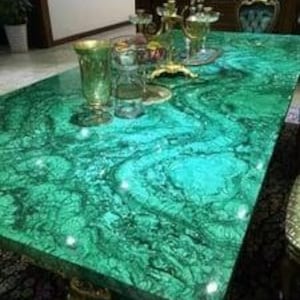 Puede incluir: Una mesa rectangular verde esmeralda con un acabado brillante. La superficie de la mesa tiene un patrón arremolinado y está adornada con detalles dorados. Varios objetos decorativos de vidrio, incluyendo copas y un jarrón, están colocados sobre la mesa.