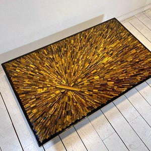 May include: Rectangular coffee table with a black frame and a mosaic top. The mosaic is made of small, rectangular pieces in shades of gold and brown, arranged in a sunburst pattern. The table sits on a white wooden floor.