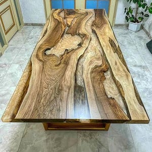 Op de afbeelding: Een rechthoekige houten tafel met een uniek ontwerp. Het tafelblad heeft een vloeiend patroon van licht en donkerbruin hout, met een heldere, glanzende afwerking. De tafel wordt ondersteund door een eenvoudige, rechthoekige houten basis. De tafel is ongeveer 180 cm lang.