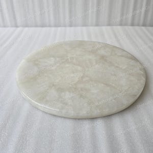 Puede incluir: Una tabla redonda de mármol blanquecino con superficie pulida. La tabla presenta un patrón veteado natural y es adecuada para servir queso, aperitivos u otras creaciones culinarias. The HandicraftsHubCo.