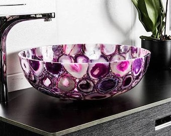 May include: A decorative, round bathroom sink with a unique agate design in shades of purple, pink, and white. The sink sits on a dark countertop next to a chrome faucet and a potted plant.