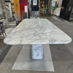 Puede incluir: Una mesa rectangular de mármol con esquinas redondeadas. La mesa presenta un patrón veteado blanco y gris. Se apoya en una base de mármol macizo y rectangular. La mesa está en un taller.
