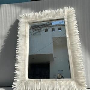 May include: Rectangular white framed mirror with a unique, textured border. The frame is adorned with numerous small, white, rectangular pieces, creating a spiky, artistic design. The mirror reflects a building and the sky.