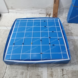 May include: A square blue display board featuring a white grid pattern. The board is encased in a matching blue frame with white stitching along the edges. Small, dark-colored rectangular markers are placed at various intersections of the grid.