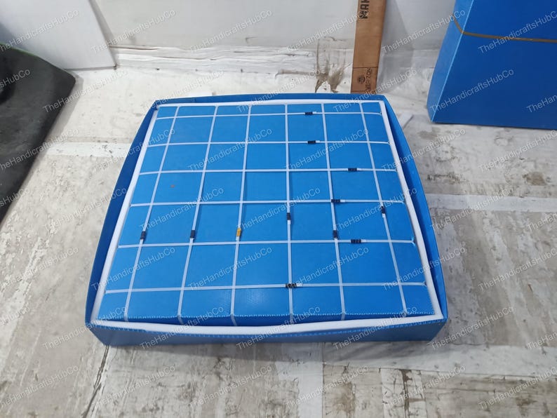 May include: A square blue display board featuring a white grid pattern. The board is encased in a matching blue frame with white stitching along the edges. Small, dark-colored rectangular markers are placed at various intersections of the grid.