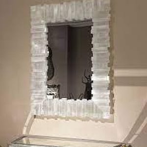 May include: A rectangular wall mirror with a textured, white frame. The frame has a layered, geometric design. The mirror is hanging on a beige wall above a mirrored cabinet.