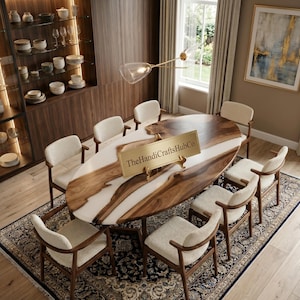 Peut inclure: Une table à manger ovale avec une surface en bois naturel et une incrustation centrale en résine blanche, entourée de huit chaises à structure en bois et assises rembourrées crème. Une plaque dorée indique TheHandiCraftsHubCo sur la table.